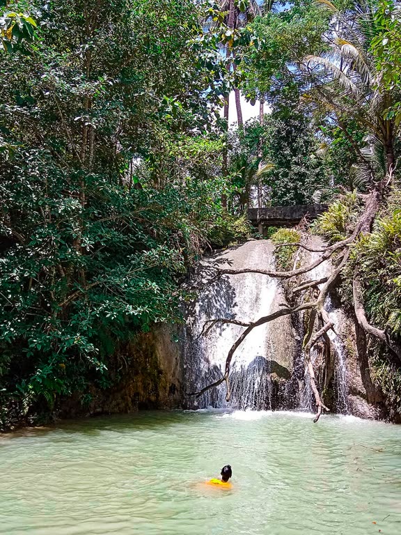 Pengunjung berenang di kolam Kedung Pengilon, dengan air terjun dan jembatan kayu di latar.