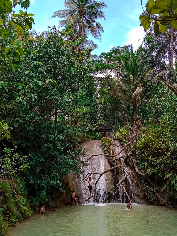 Air terjun Kedung Pengilon dan kolamnya dilihat dari tepi, dengan pohon kelapa dan langit biru.